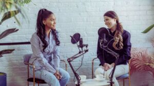 Two women recording a podcast about autism support and ABA therapy, seated in a modern setting with microphones and coffee cups. The image shows their engaging process of creating BCBA podcasts that include discussions for families and ABA professionals.