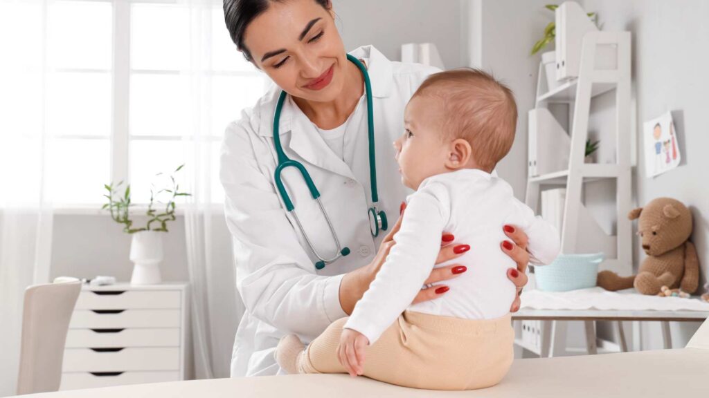 A baby sitting on an exam table with a doctor nearby, illustrating a routine visit about 12‑month‑old developmental milestones