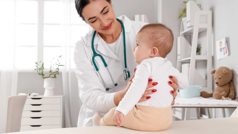 A baby sitting on an exam table with a doctor nearby, illustrating a routine visit about 12‑month‑old developmental milestones