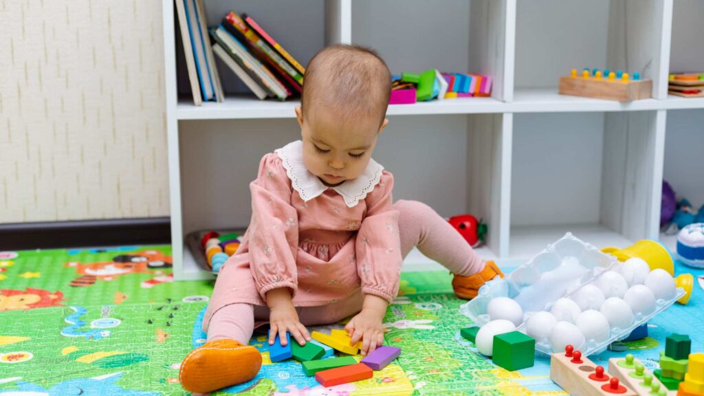 A baby sitting on the floor playing with toys, used to illustrate early signs of autism at 18 months