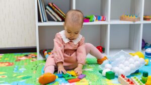 A baby sitting on the floor playing with toys, used to illustrate early signs of autism at 18 months