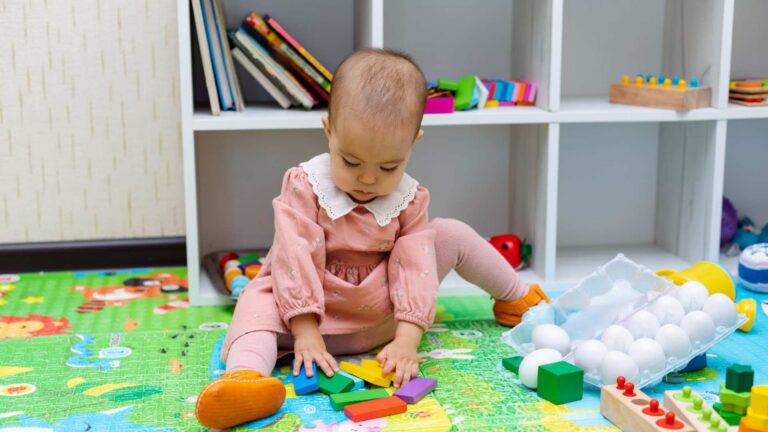 A baby sitting on the floor playing with toys, used to illustrate early signs of autism at 18 months