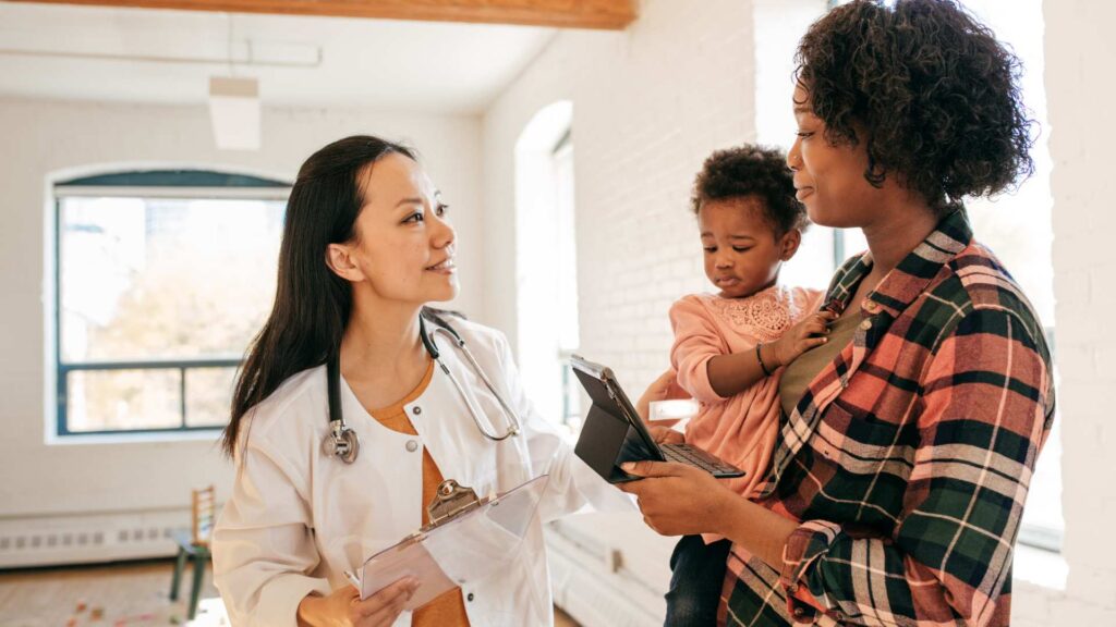 A pediatrician reviews the M-CHAT screening questionnaire with a mother holding her toddler during a well-child visit