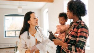 A pediatrician reviews the M-CHAT screening questionnaire with a mother holding her toddler during a well-child visit