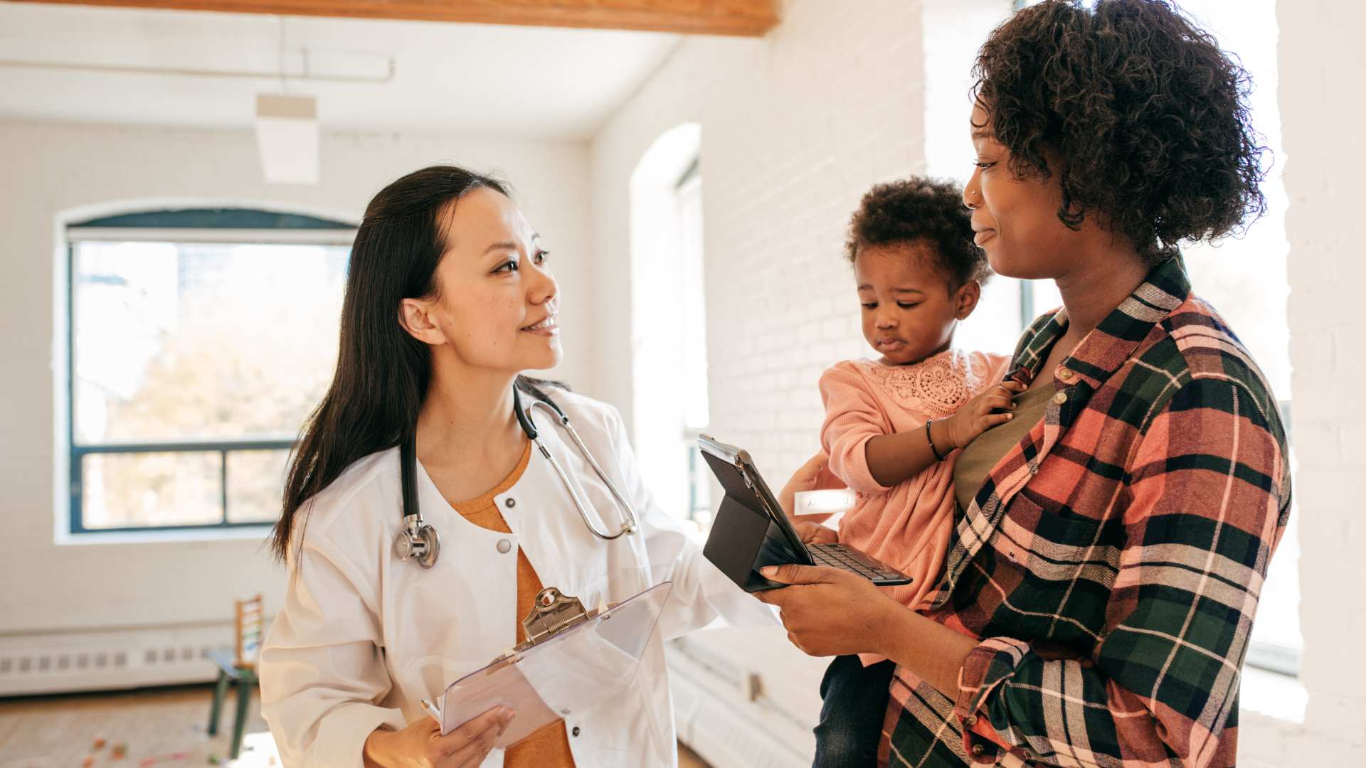 A pediatrician reviews the M-CHAT screening questionnaire with a mother holding her toddler during a well-child visit
