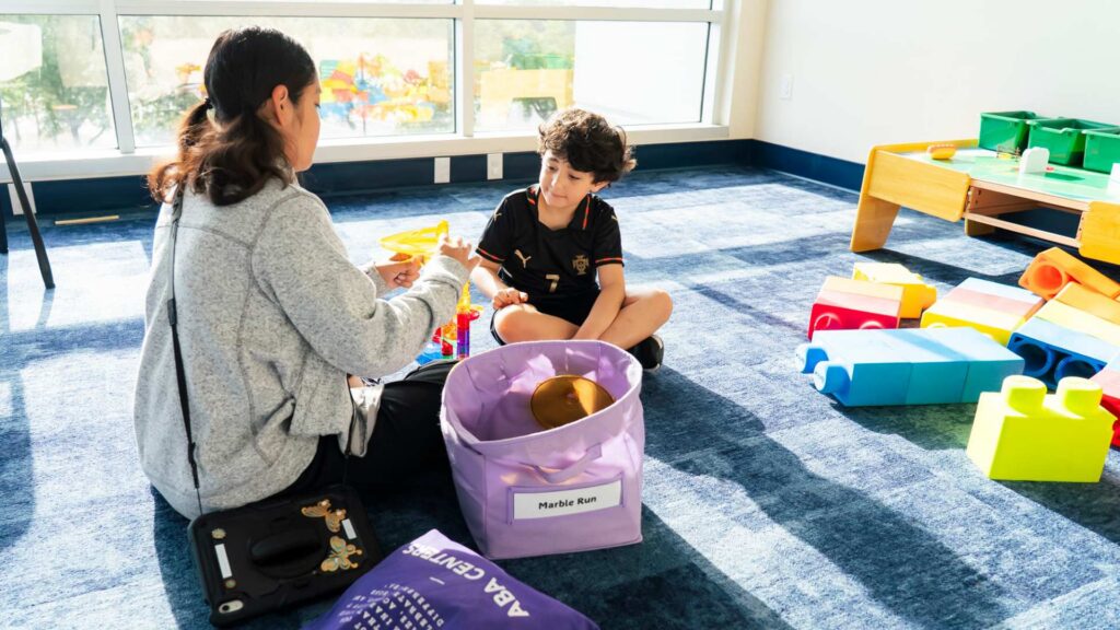 ABA therapist and a boy sit on the floor sorting toys into bins surrounded by colorful building blocks during a play-based ABA therapy session