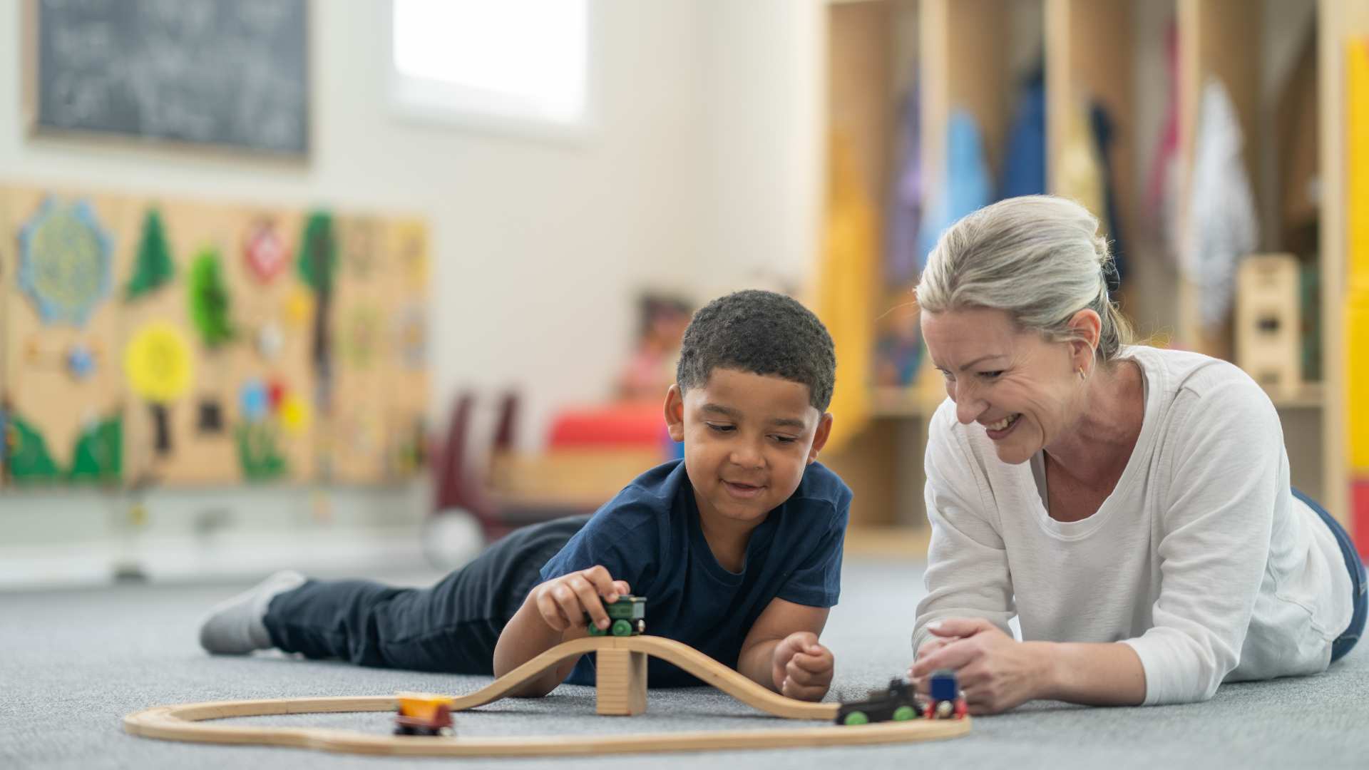 ABA therapist and a young boy lie on the floor playing together with a wooden train set during a play-based ABA therapy session