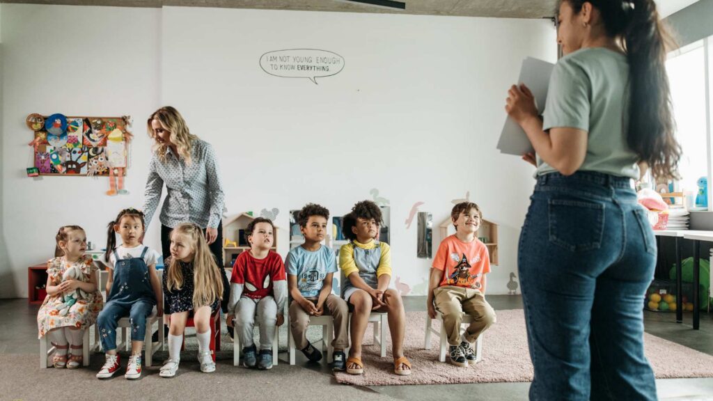 ABA therapist holds a clipboard while leading a group session with young children seated in a school classroom, supported by their teacher