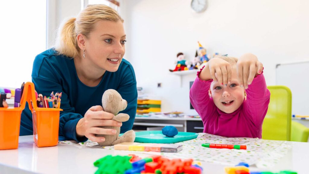 ABA therapist holds a stuffed animal while a toddler girl plays with modeling clay during an ABA therapy for toddlers in a colorful playroom