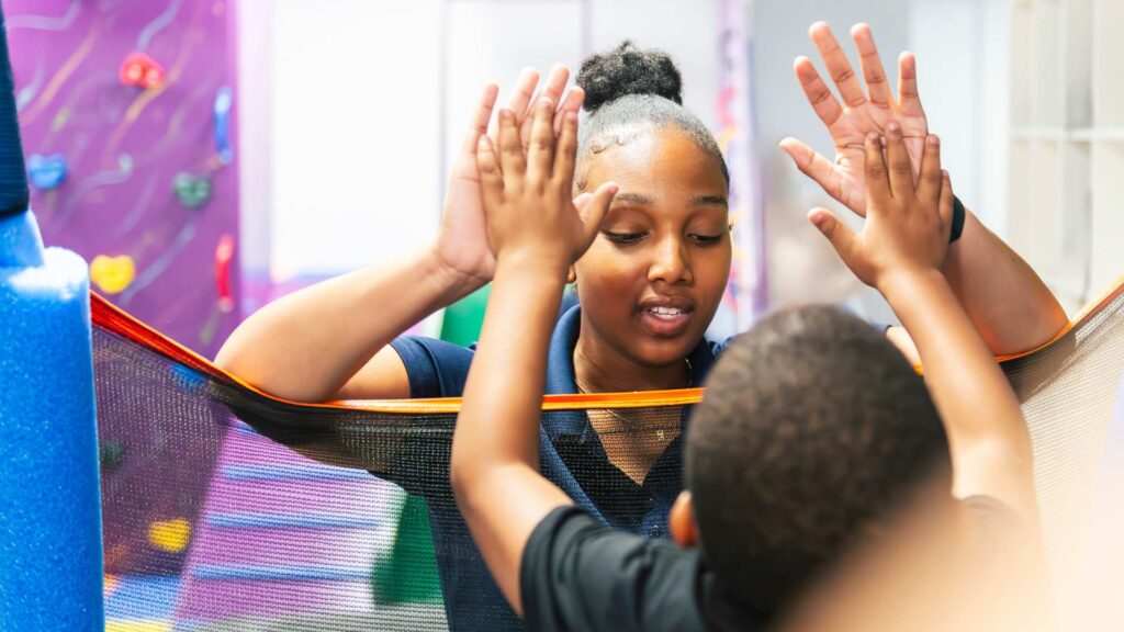 ABA therapist raises her hands during an interactive activity with a young boy in a colorful sensory play area during an ABA therapy session