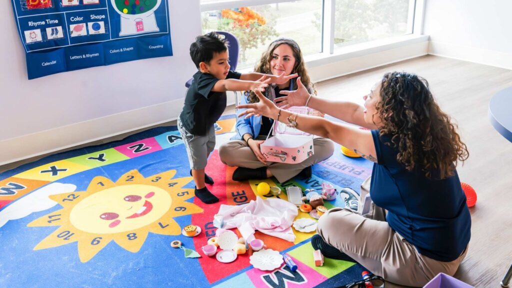ABA therapy provider and a therapist engage a young boy in a playful hand-gesture activity on a colorful alphabet rug in a bright clinic room
