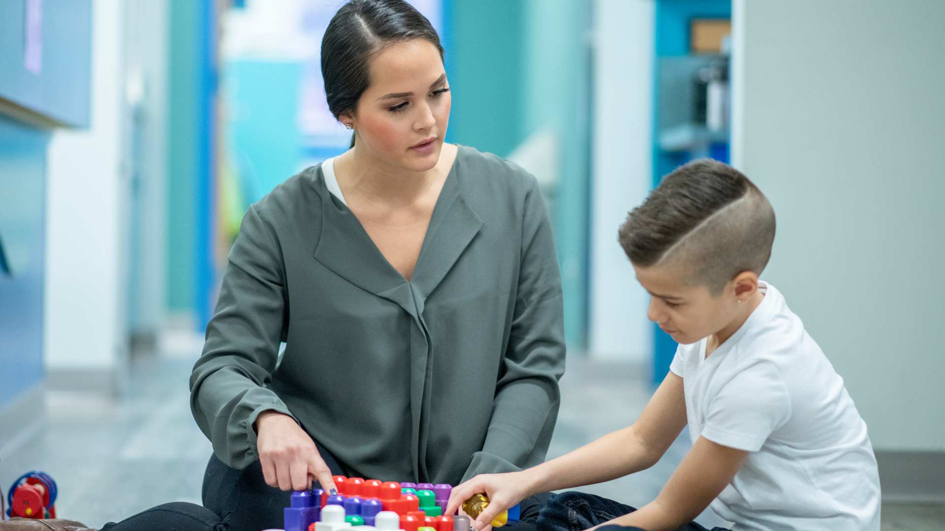 ABA therapy provider sits on the floor with a young boy, guiding him through a building block activity during a clinic session