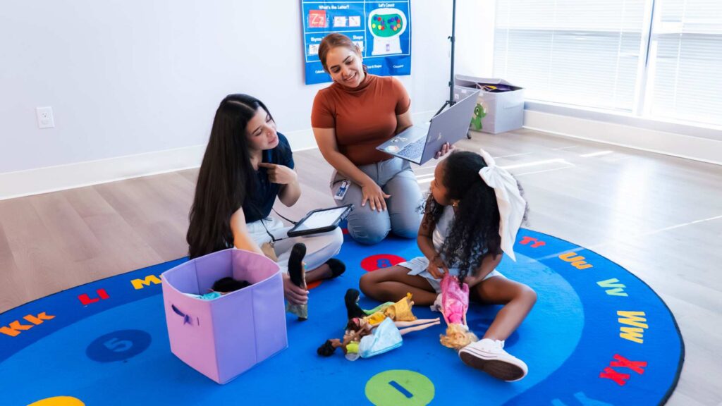 BCBA observing an ABA therapy session while a therapist interacts with a young girl playing with dolls on a colorful alphabet rug in a clinic room