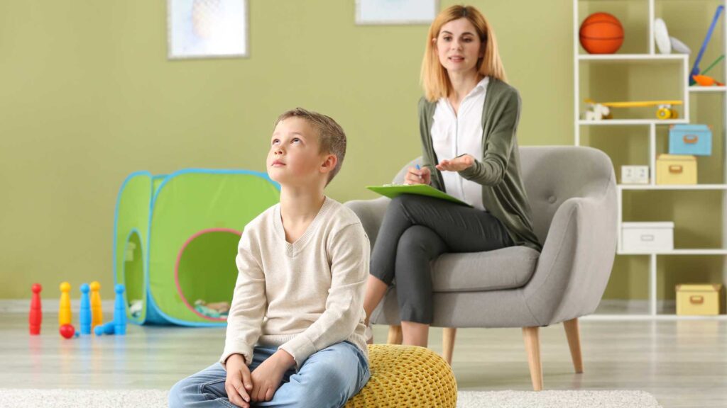 Autism therapy: A child sitting on a beanbag chair in a room with a woman holding a clipboard.