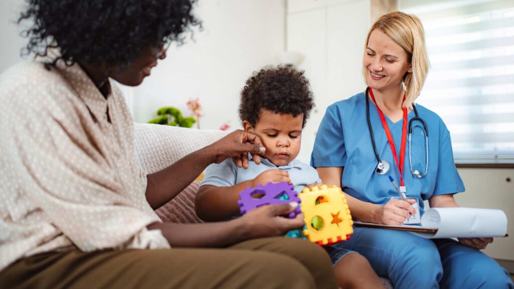 Healthcare professional takes notes on a clipboard while observing a toddler playing with colorful shape blocks beside his mother during an autism evaluation report visit