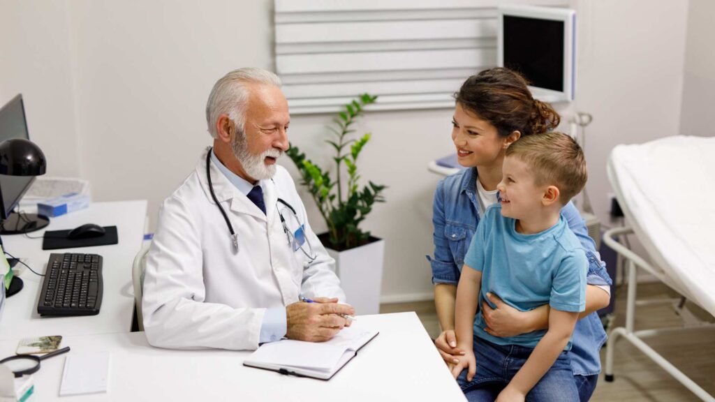 Mother holds her young son on her lap while a smiling pediatrician speaks with them during a clinic appointment