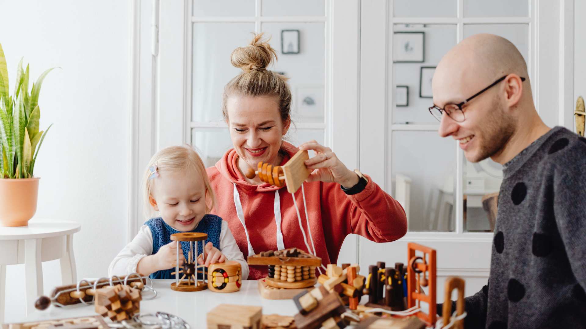 Parents and their toddler daughter smile while playing together with wooden developmental toys at a table, illustrating the role of parents in ABA therapy