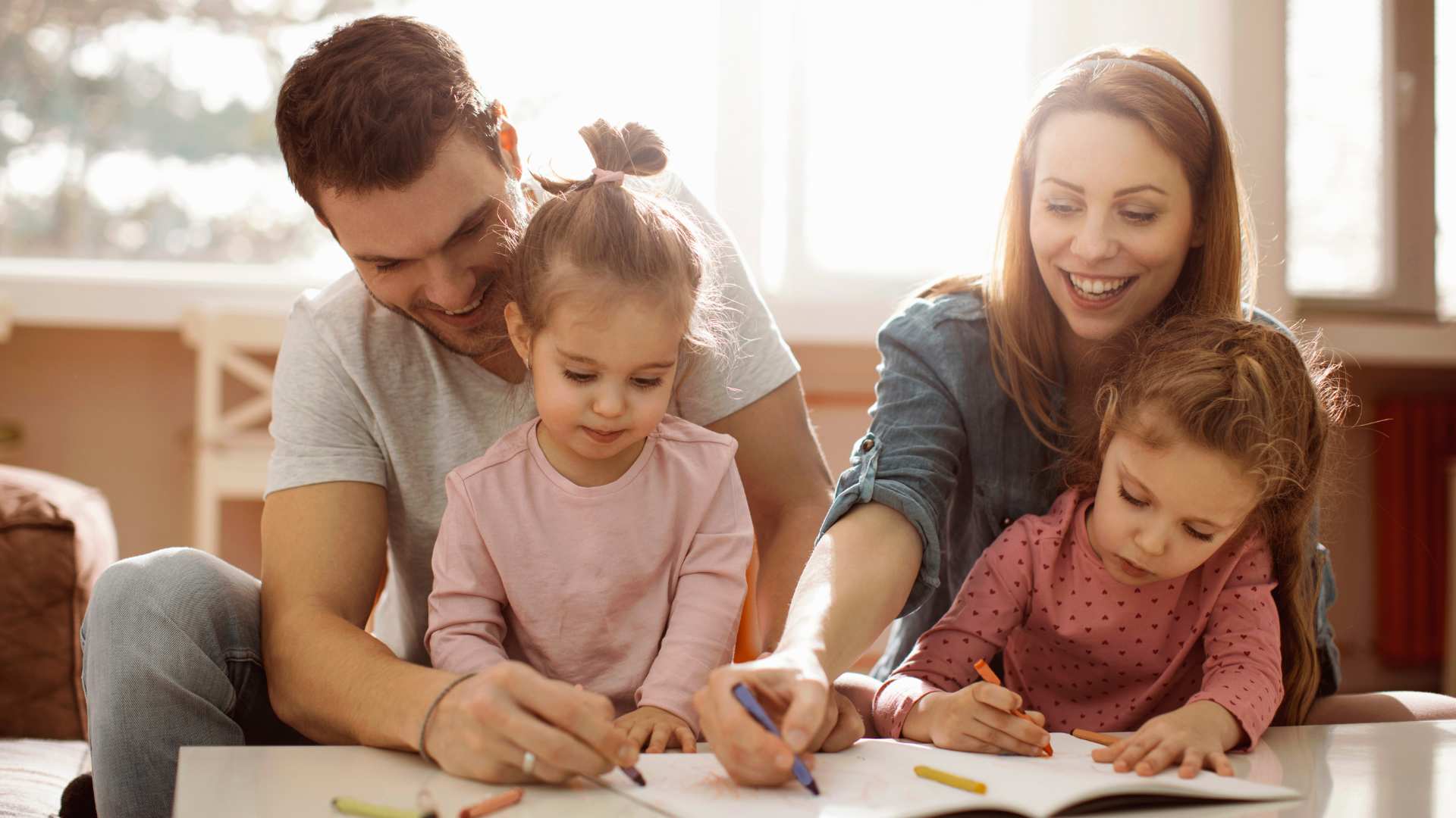 Parents sit with their two young daughters coloring together with crayons at a table, showing parental involvement in ABA therapy activities at home