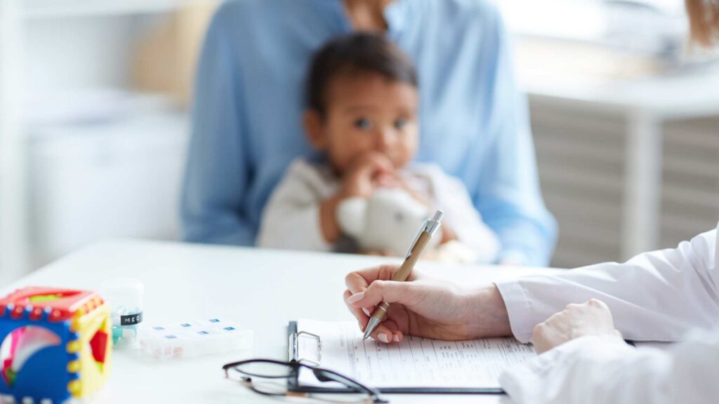 Pediatrician writes notes on an evaluation form while a mother holds her toddler in the background during an autism assessment appointment