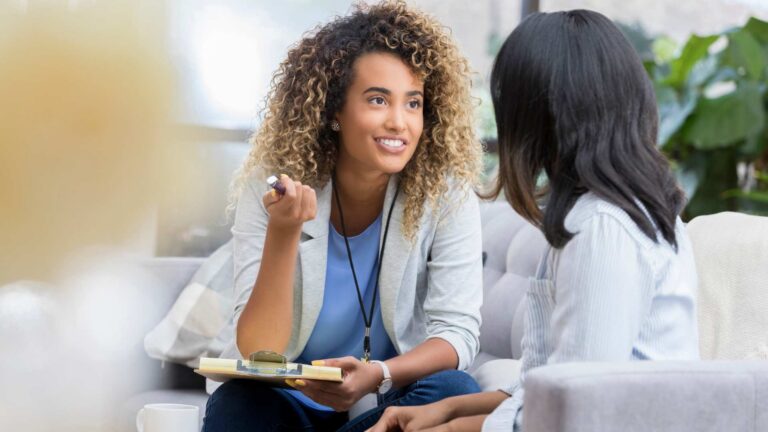 Qualified ABA therapist with a clipboard gestures while speaking to a client on a couch during a therapy session, highlighting ABA therapist qualifications in practice