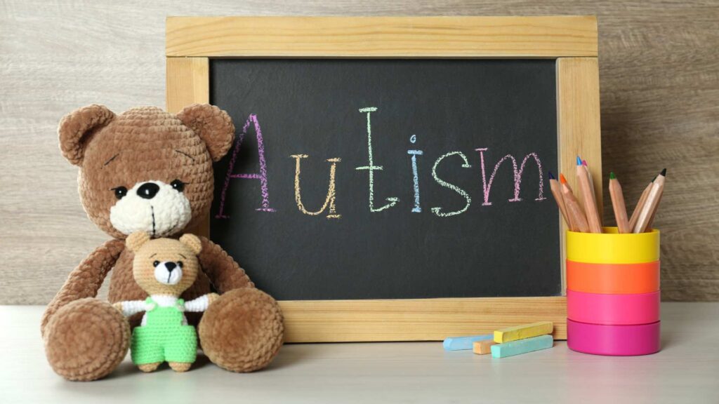 Small blackboard with the word 'Autism' written in colorful chalk, surrounded by stuffed bears, colored pencils, and chalk pieces on a wooden table