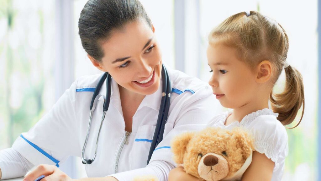 Smiling pediatrician interacts with a little girl holding a teddy bear during an autism assessment appointment