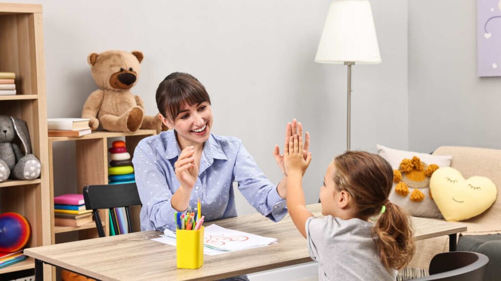 Smiling psychologist gives a high five to a little girl at a table with crayons and drawings during an autism evaluation session