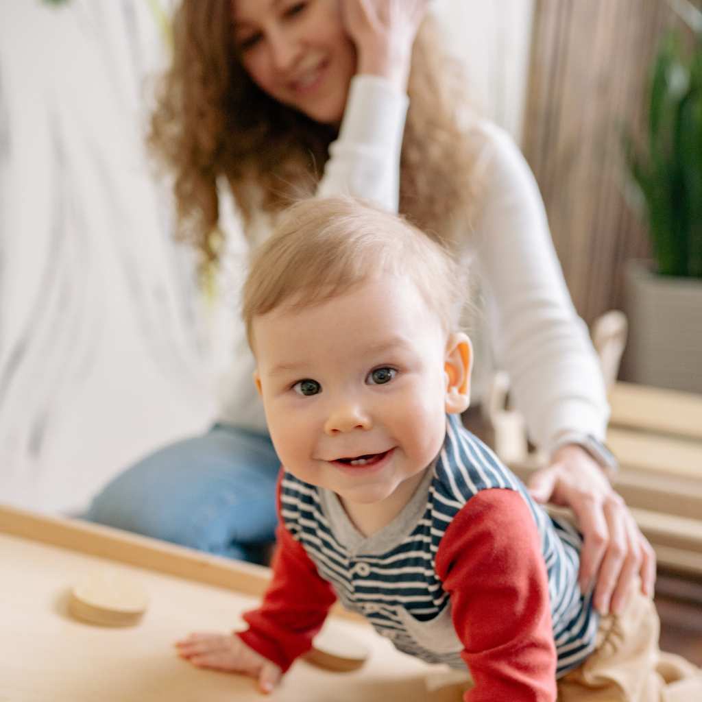 Smiling toddler crawling on a wooden surface with an adult behind them