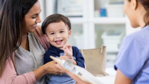 Smiling toddler sits on his mother's lap while a pediatrician holds a clipboard during an autism evaluation appointment