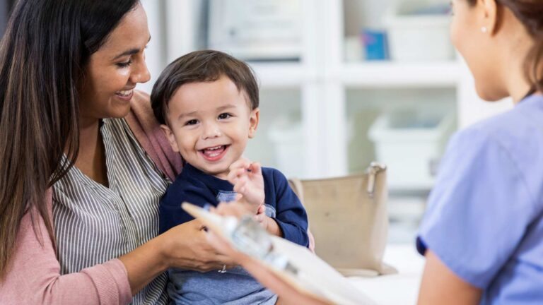 Smiling toddler sits on his mother's lap while a pediatrician holds a clipboard during an autism evaluation appointment