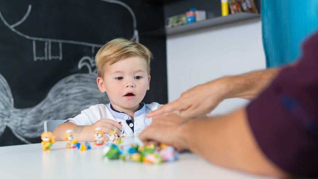 Toddler boy engages with small toy figures on a table while a therapist guides him during an early intervention session in a clinic with a chalkboard behind them
