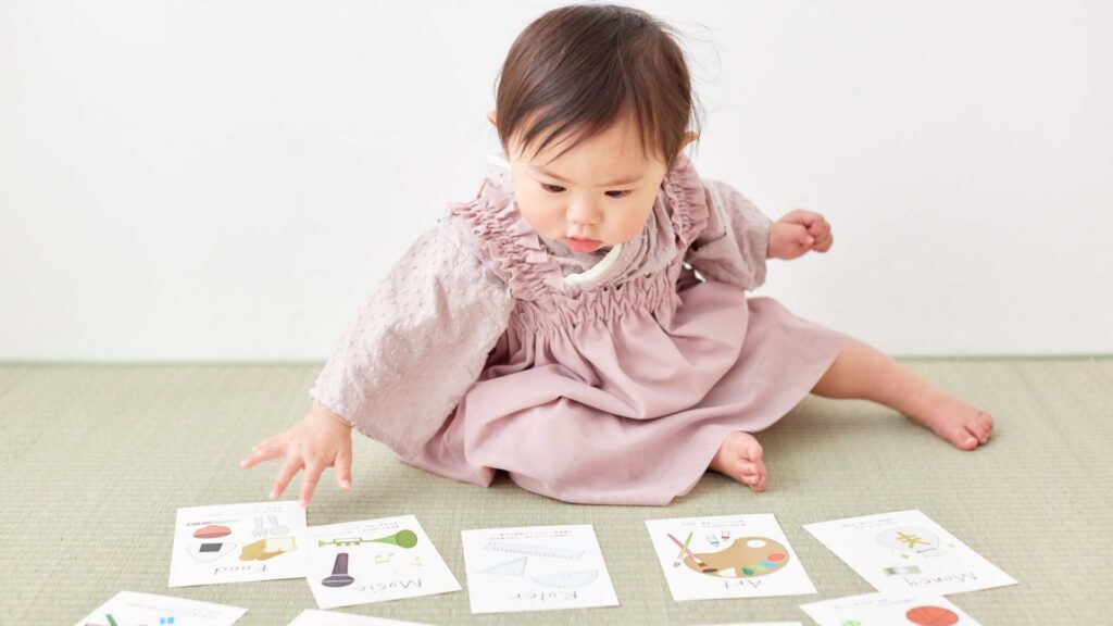 Toddler in a pink dress reaches for educational flashcards spread on the floor during an early intervention learning activity