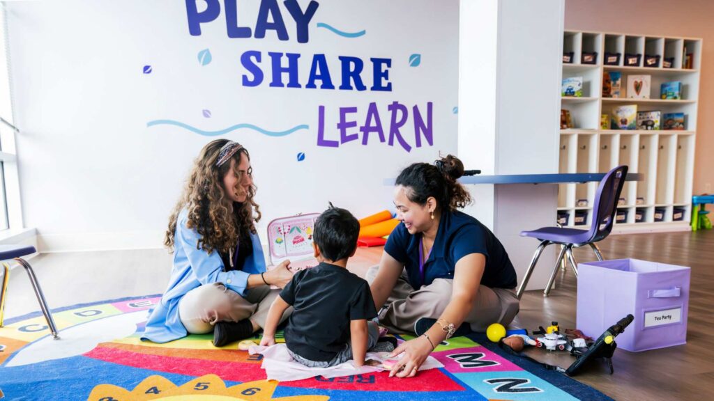 Two ABA therapists sit on a colorful alphabet mat with a young boy during a play-based therapy session in a bright classroom with a 'Play, Share, Learn' sign on the wall