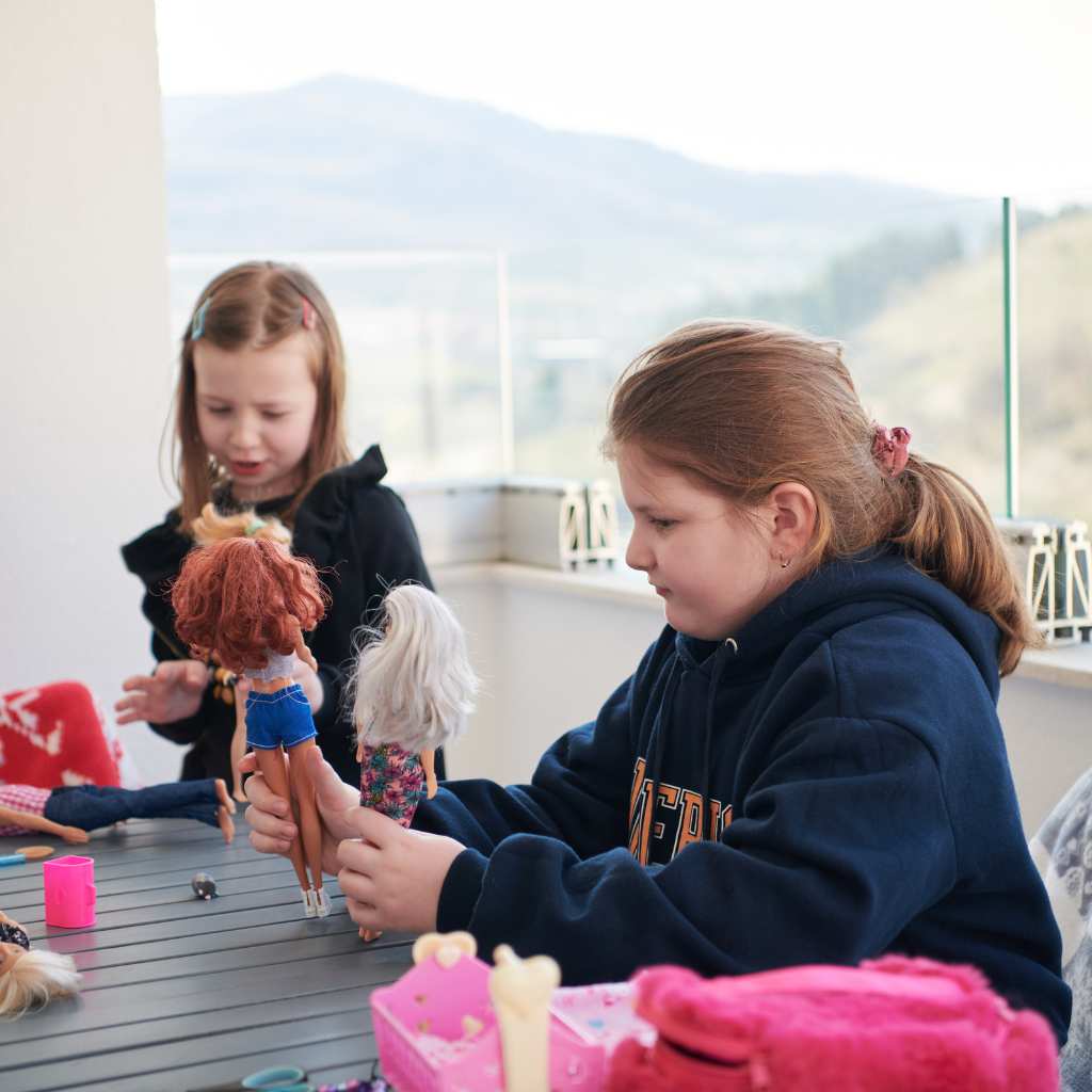 Two children playing with dolls at an outdoor table