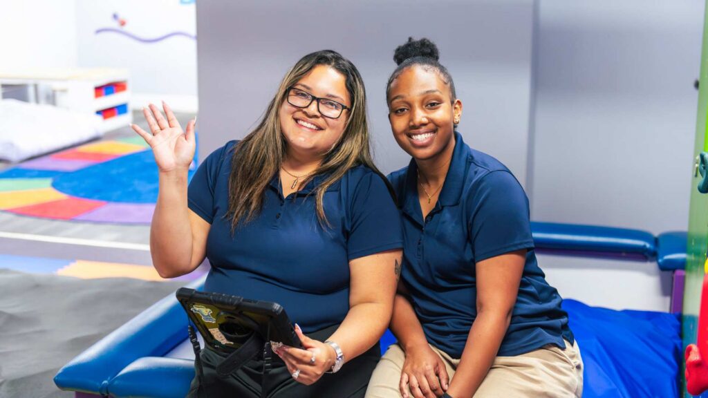 Two smiling ABA professionals in navy polo shirts sit together in a colorful therapy clinic, one waving while holding a tablet