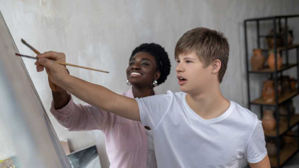 Young neurodivergent person painting with support, symbolizing the paradigm shift from deficits to the neurodiversity model that values different ways of thinking