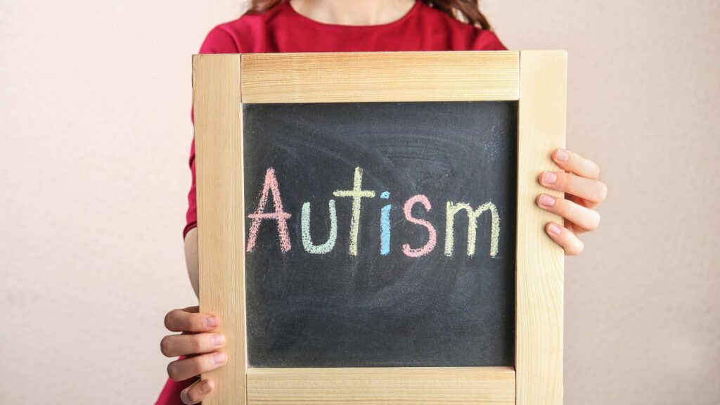 Woman in a red sweater holds a chalkboard with the word 'Autism' written in colorful chalk letters against a light background.