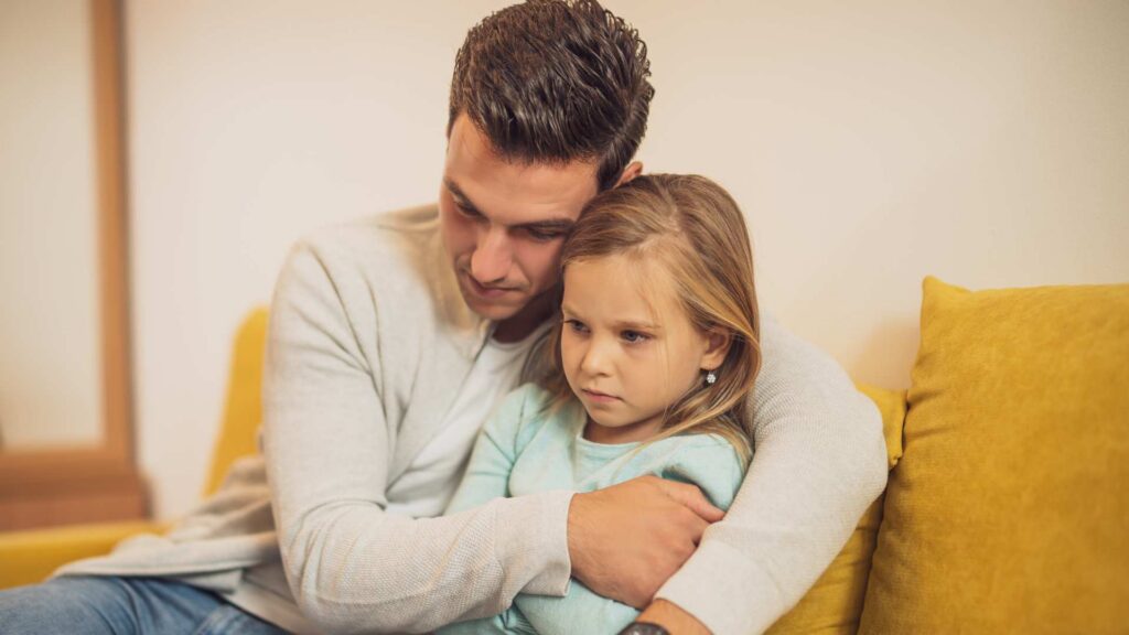 Worried father holds his young daughter on a yellow couch, both looking concerned after an autism diagnosis