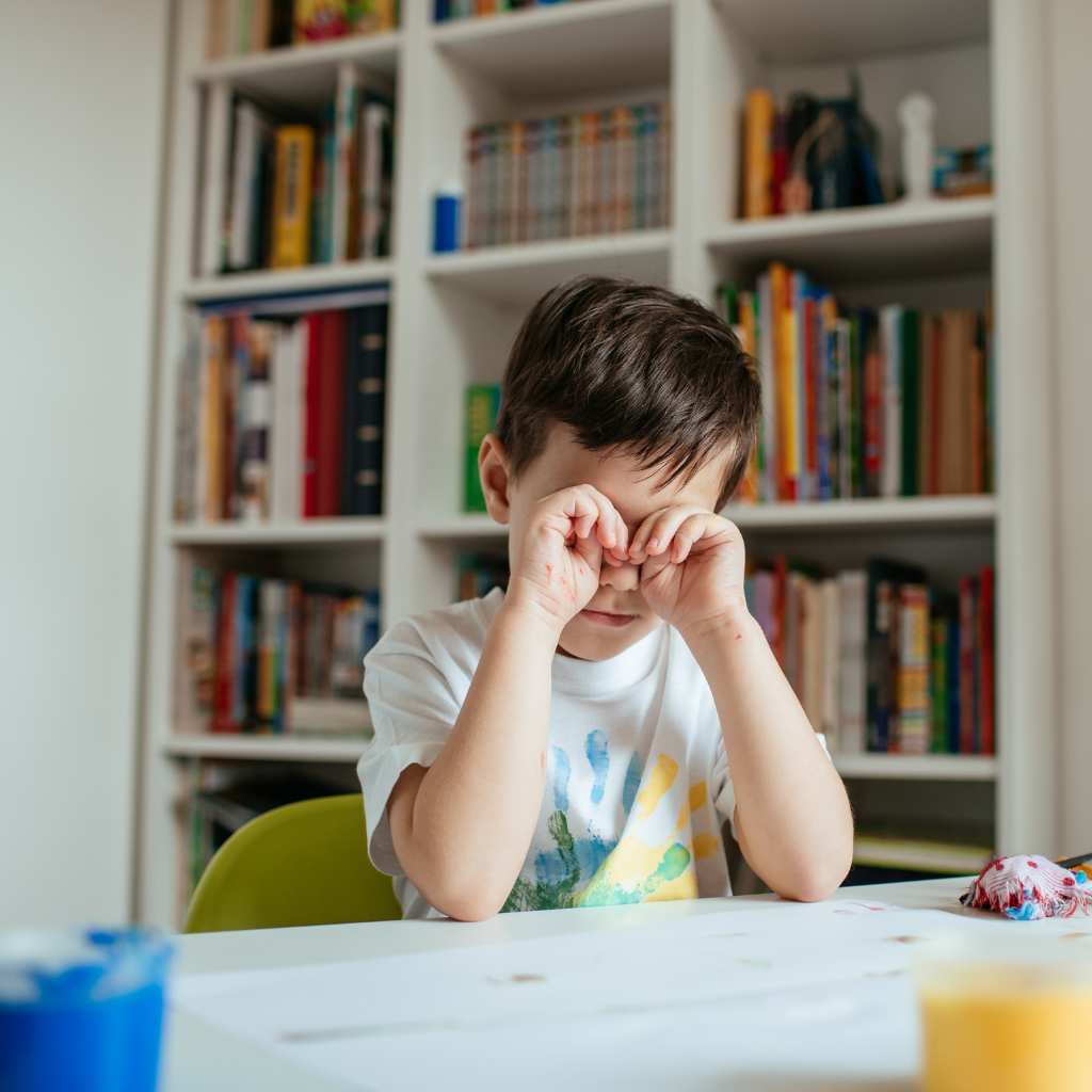 Young boy rubbing his eyes at a table after a painting activity, showing signs of fatigue or sensory overwhelm