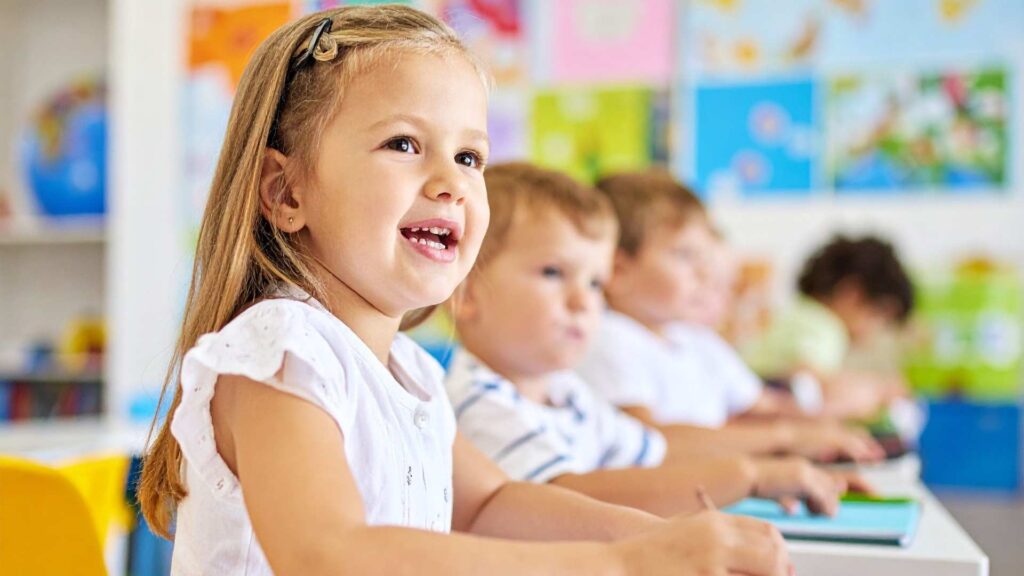 Young child smiling at a desk in a lively classroom, capturing a calm moment connected to cooperation skills in autism