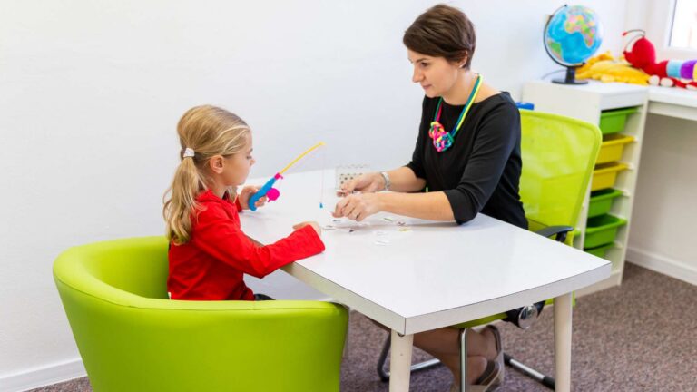 Young girl uses a toy fishing rod during a playful therapy activity with a BCBA at a table in a colorful clinic room. What is a BCBA