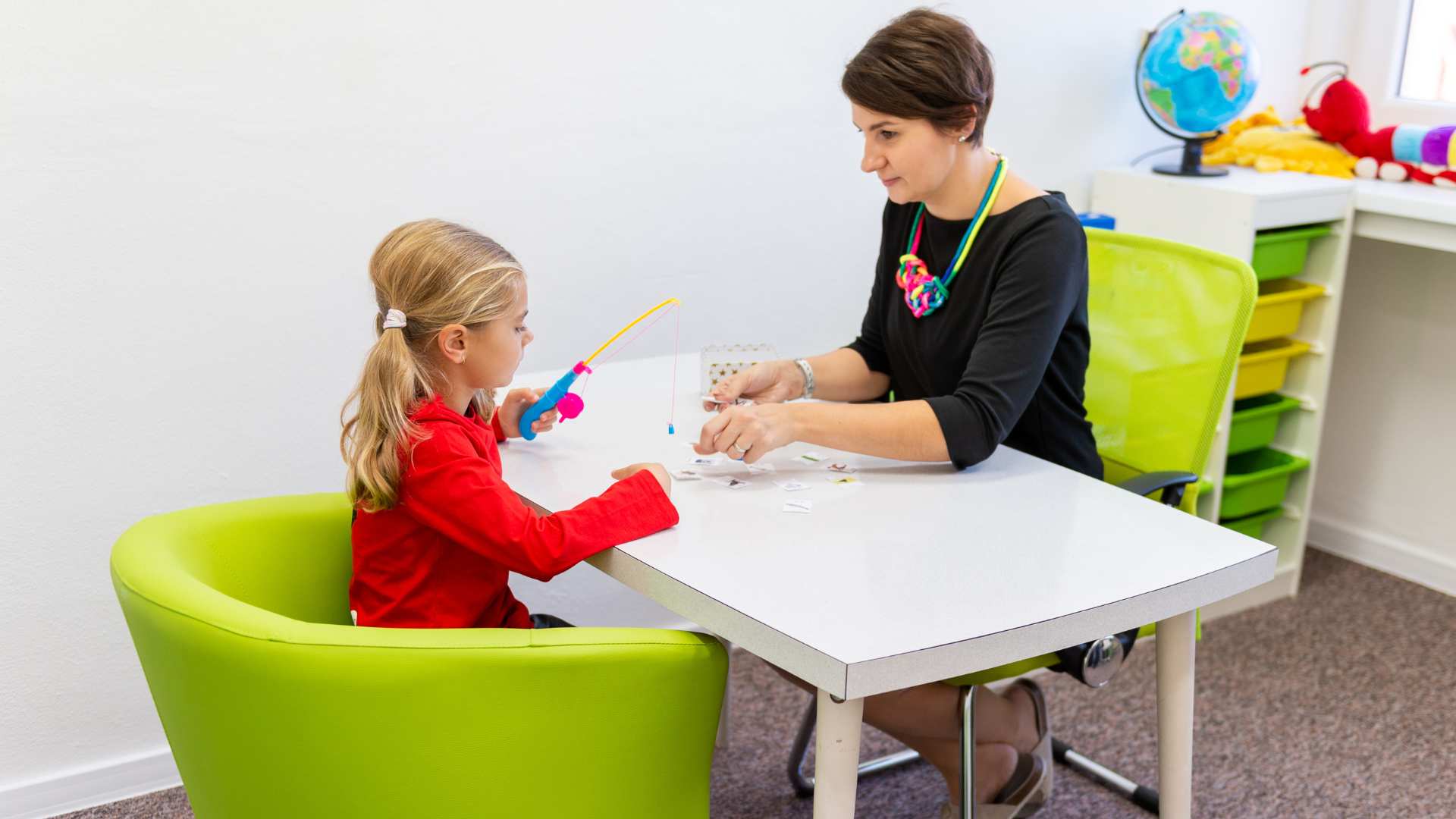 Young girl uses a toy fishing rod during a playful therapy activity with a BCBA at a table in a colorful clinic room. What is a BCBA